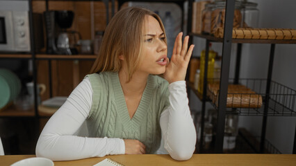 Young blonde woman cups hand over mouth calling out across wooden kitchen table next to shelving...