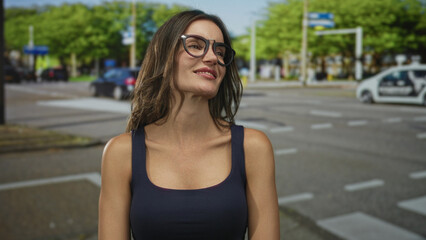 Woman in casual navy tanktop and oversized glasses stands smiling on sunlit city street sidewalk with blurred cars; joy.