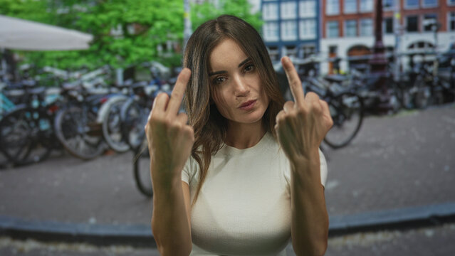 Young hispanic woman in white t-shirt flips middle fingers with bare hands on city street; anger defiance.