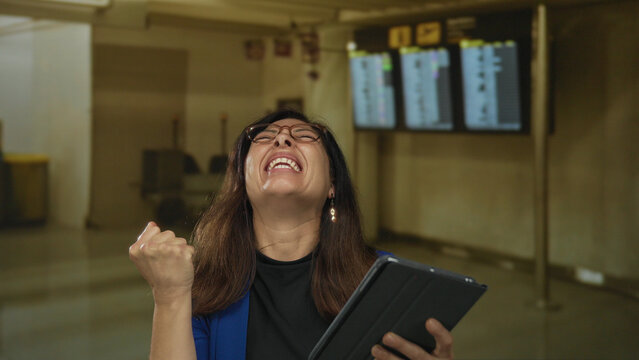 Woman in blue blazer and glasses holds tablet and pumps fist in crowded airport terminal while smiling broadly with excitement; victory.