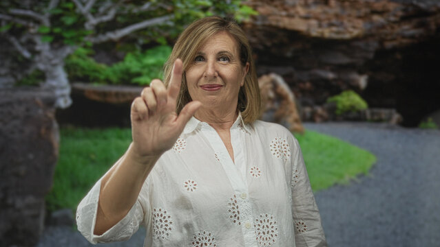 Woman holds up open hand palm forward in green forest park with tree trunks and mossy rock path; serenity.