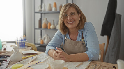 Woman folded hands at ceramics table inside art studio surrounded by clay tools, pottery vases and paintbrushes; contentment.