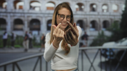 Fototapeta premium Woman making finger heart gesture with fingers and smiling in front of the roman coliseum building, casual outfit visible; playful travel.