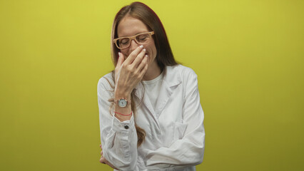 Fototapeta premium Scientist woman in white lab coat covering mouth with hand and wearing glasses in yellow studio; joy playful warmth.