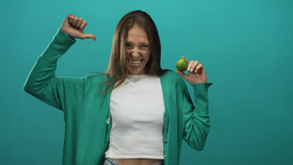 Woman holding a lime and giving thumbs down in a teal studio while wearing a green cardigan and...