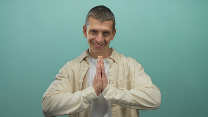 Man smiling with hands in prayer gesture against blue wall wearing casual shirt isolated in studio...