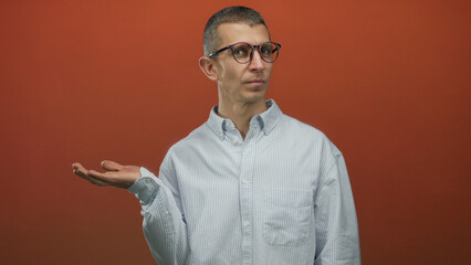 Man gestures with one hand while pointing with a finger against a red wall, wearing glasses and a light shirt, suggesting confusion or explanation in an isolated setting.