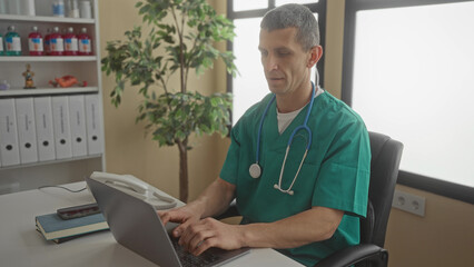 Doctor in green uniform with stethoscope using laptop in a hospital clinic office with organized files and plants indoors.
