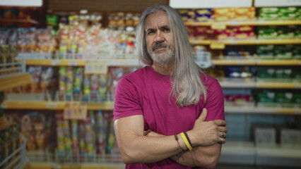 Man with long gray hair wearing magenta shirt holds forearm beside stocked supermarket shelves in building aisle; contemplation resilience aging everyday.