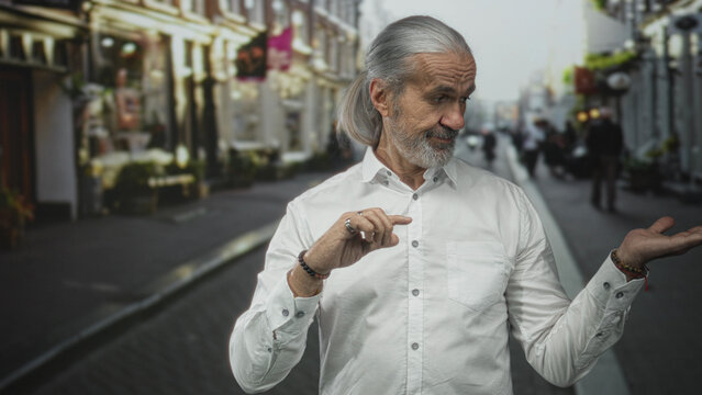 Fototapeta Man points finger with right hand and shows open left palm while standing on a busy city street in daylight, wearing white shirt  subtle confidence wisdom.