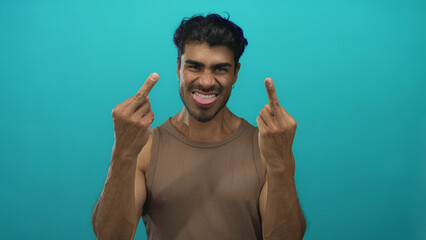Man with raised hands flips both middle fingers and sticks out tongue in a turquoise studio;...
