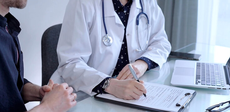 Doctor and patient are talking in clinic after usual health check up. Female physician is making notes awhile man is describing something. Medicine concept