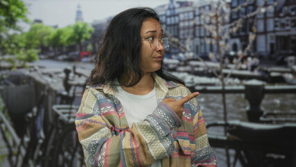 Fototapeta premium Woman pointing finger while shrugging hands near canal boats and bicycles on street in amsterdam; skeptical curiosity.
