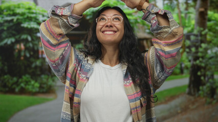 Woman with glasses smiles and raises hands above head while standing on a leafy forest path wearing a white tee and patterned jacket; joy wonder.