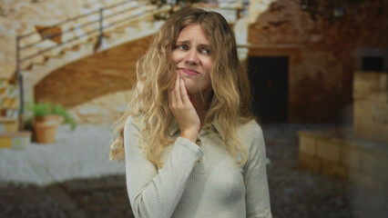 Portrait captures woman with tooth discomfort, grimacing against a stone courtyard wall as a blonde...