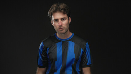 Naklejka premium Man smiling with slight head tilt wearing blue and black soccer jersey in studio against black backdrop; confidence.
