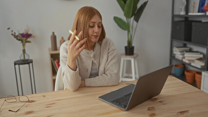 Woman contemplating indoors with laptop and holding a pen, seated at wooden table in modern apartment living room with bookshelves and houseplants.