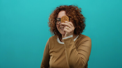 Woman holding cookie with happy expression against blue backdrop, showcasing middle-aged hispanic woman enjoying snack in isolated setting.