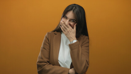 Hispanic woman laughing warmly against an orange wall, covering her mouth with her hand, wearing a...