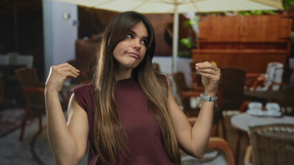Woman gesturing on an outdoor terrace at a coffee shop in a lively city setting, showing expressive hand movements typical in hispanic culture with tables and chairs in the background.