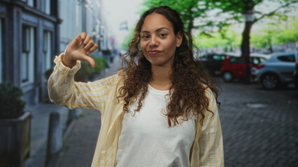 Fototapeta premium Young african american woman showing thumbs down with hand raised on city street near parked cars and trees, casual shirt and neutral expression; disapproval rejection critique.