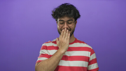 Young man with beard and glasses smiles gently and covers his mouth against a vibrant purple background in casual red striped shirt suggesting amusement and candid emotion.