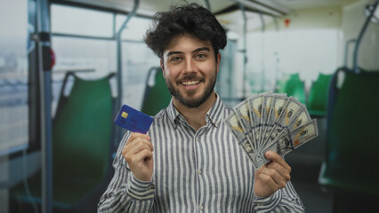Young man with beard examines credit card and holds fan of cash while sitting on a bus indoors,...