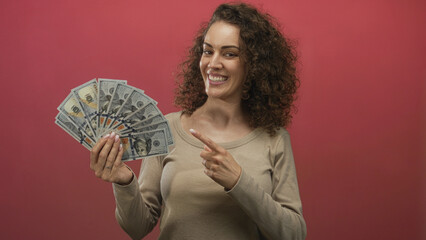 Young hispanic woman holding a fan of us dollar bills with visible hand and pointing finger toward...