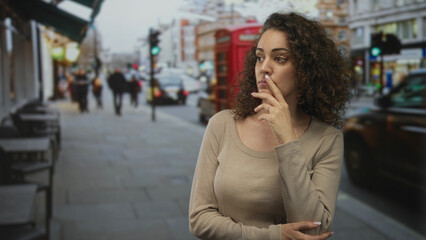 Obraz premium Woman wearing beige sweater finger to lips on a busy city street near a red phonebox and taxi; thoughtful uncertainty.