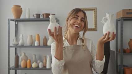 Young blonde woman wearing apron holds two handcrafted ceramic vases in pottery studio; creativity pride joy.