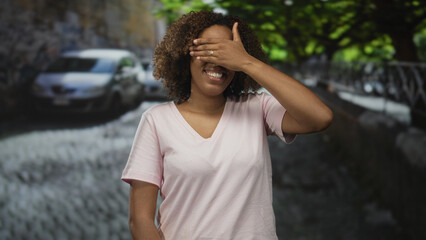 Woman covers eyes with hand and smiles on a cobblestone street wearing a pink t shirt; playful...