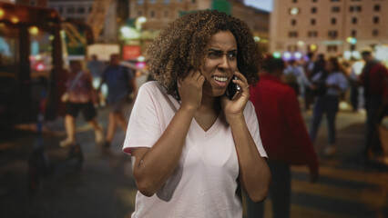 Woman covering ears while holding smartphone and wincing on a crowded city street; frustration noisy call disruption.