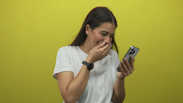 Young brunette woman tapping smartphone screen and holding phone while smiling, wearing white tshirt and smartwatch in yellow studio wall; lighthearted joy.