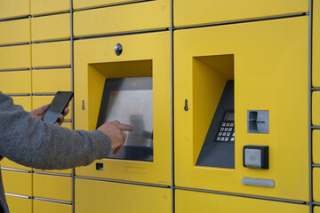 Man using smartphone and touchscreen to open yellow parcel locker for online delivery pickup