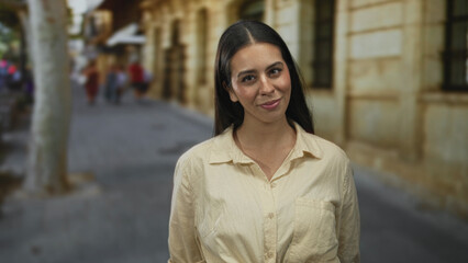 Woman smiling with visible face and open collar, wearing beige shirt, standing on a sunny city street near stone building and blurred pedestrians; gentle joy.