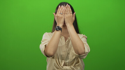 Woman with raised hands covering face and sticking out tongue in studio with green backdrop;...