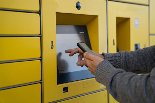 Person using smartphone and touchscreen terminal on yellow parcel locker for online delivery pickup