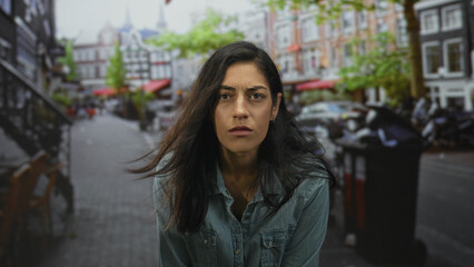 Woman frowning face, hair windblown, wearing denim shirt, leaning forward and staring at camera on a city street with cafes and cobblestones; frustration resilience.