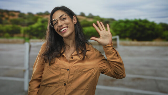 Woman waving right hand on outdoor tennis court near net, wearing glasses and orange shirt, smiling at camera; friendly greeting joy.