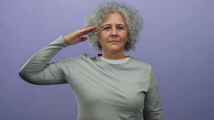 Woman with grey hair saluting against isolated purple wall wearing casual grey attire.