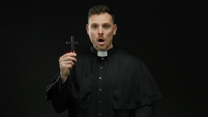 Man dressed as priest holding a cross against a dark background with a hand over his mouth, conveying surprise or shock, isolated and mysterious religious imagery in silhouette.