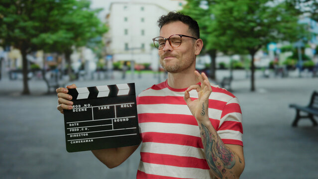 Handsome man smiling on city street holding film clapperboard wearing striped shirt glasses and tattoos urban background creating vibrant outdoor scene.