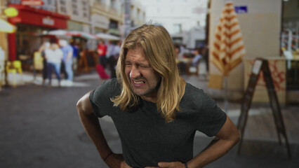 Blond man with long hair visibly grimaces as he clutches stomach on street; pain discomfort nausea distress.