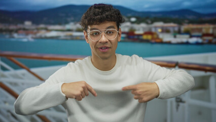 Fototapeta premium Young hispanic man wearing glasses and sweater waving hand on cruise deck with railing and sea in view; adventure greeting travel excitement.