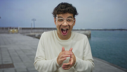 Man clapping hands on street wearing white sweater and eyeglasses beside waterfront promenade under...