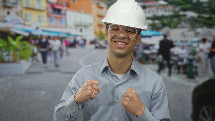 Hispanic engineer man wearing hardhat clenching fists and smiling on a busy city street; optimism...