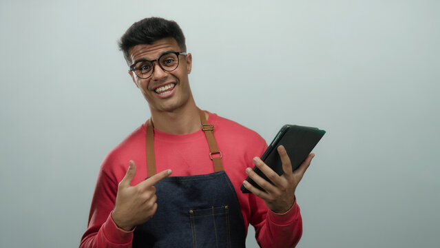 Young man wearing glasses and apron smiles while holding tablet and giving thumbs up against white background