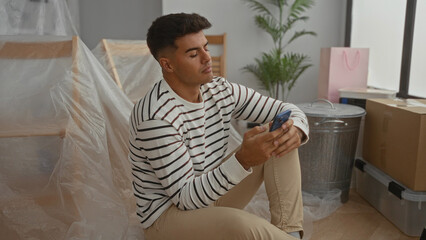 Young man sitting indoors in new home surrounded by moving boxes using smartphone looking thoughtful and relaxed