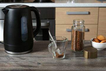 A glass cup, an electric kettle and a jar of instant coffee on the table against the background of the interior of a home kitchen.