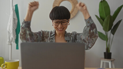 Young woman with glasses working on a laptop at home, celebrating success in a cozy living room with plants and modern decor.